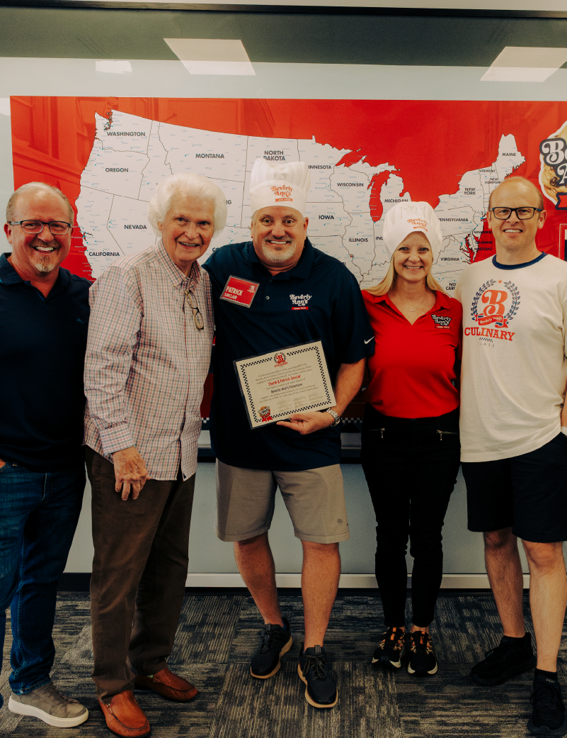 Four Travelin’ Tom’s Coffee Truck franchise owners stand in front of a map of the U.S. while one person points to their franchise’s location