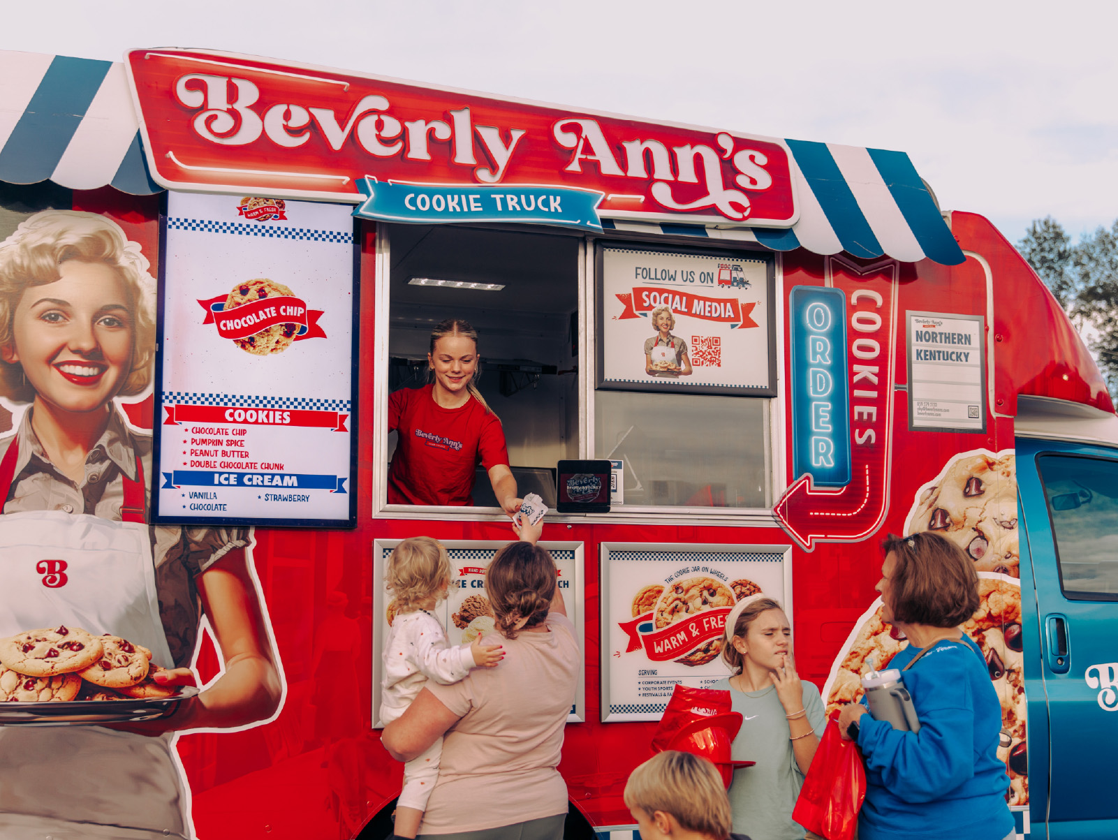 A Travelin’ Tom’s Coffee Truck franchisee hands a customer a beverage through a window in her Beverage Entertainment Vehicle, while customers stand in line.